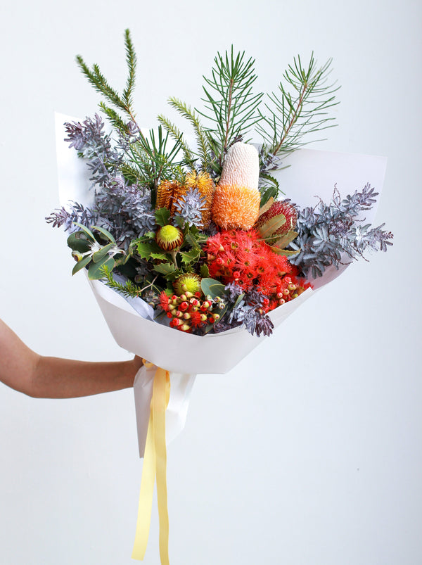 A woman is holding a large Native style bouquet of flowers. The bouquet is wrapped in white paper and is tied with a pastel yellow ribbon. The bouquet features a variety of Banksias, red flowering gum and Dryandra