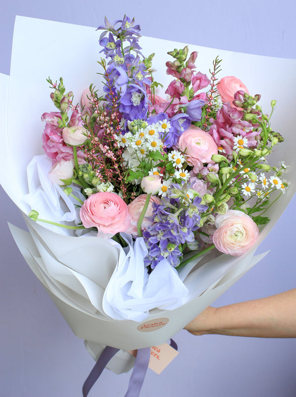 A pastel toned bouquet of seasonal garden style flowers. The flowers are wrapped in white paper and are held in front of a lilac coloured backdrop.