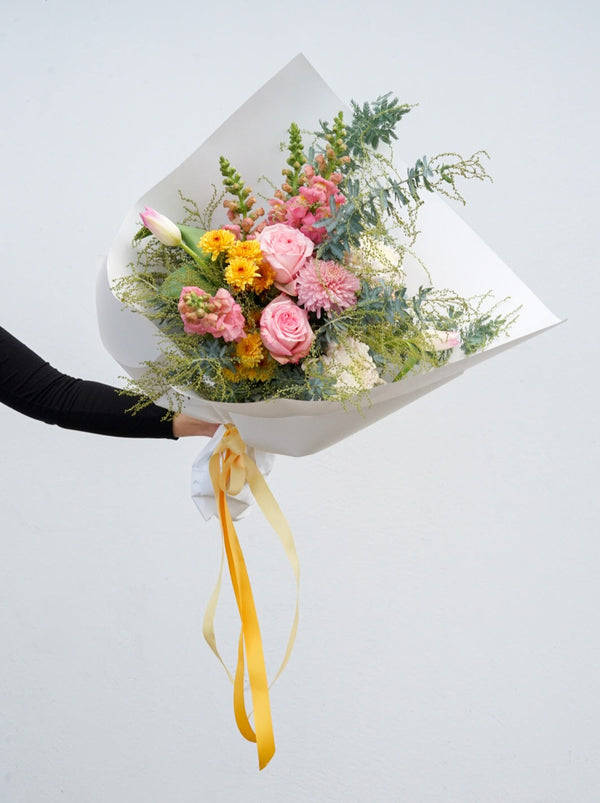 A pink and yellow bouquet of roses, tulips, wattle, snapdragons and chrysanthemum. The bouquet is gift wrapped in white paper and tied with a yellow ribbon for delivery