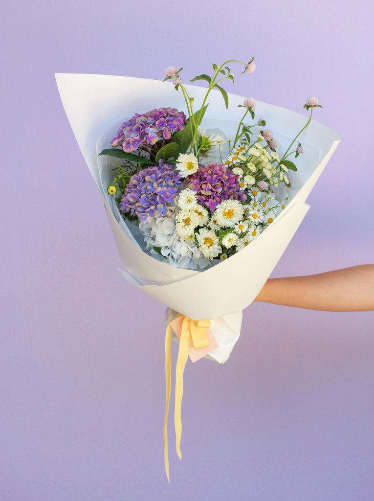 A small bouquet of flowers gift wrapped in white paper and finished with a pastel yellow ribbon. The flowers in the bouquet are purple and white hydrangea, matricaria chamomile daisies, lilac coloured gomphrema and cosmos. The bouquet is held in an outstretched arm in front of a lilac background. 